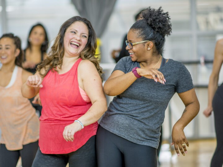 A multi-ethnic group of adult women are dancing in a fitness studio. They are wearing athletic clothes. Two women are laughing while dancing together.